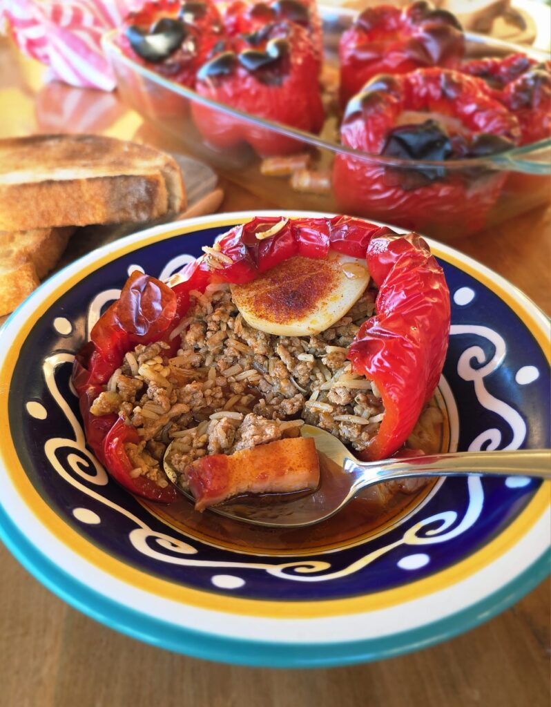 Close-up photo of a similar dish, Balkan Stuffed Capsicums (Punjena paprika) that has a similar filling to the current recipe, Balkan Stuffed Cabbage Leaves (Sarma). Served in a decorative blue and white bowl, and cut open to show the pork mince filling inside. 