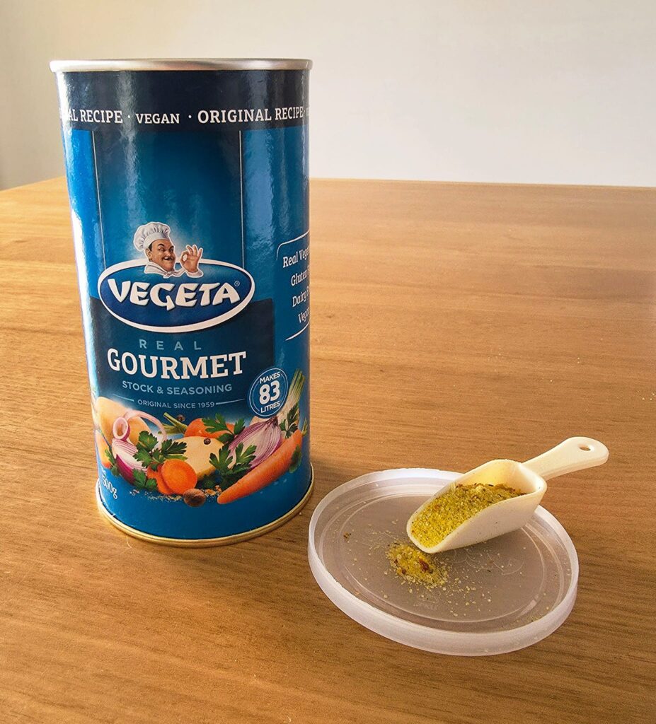 Close-up photo of a container of Vegeta stock powder/seasoning, with a small white scoop showing some of the powder, all set on a wooden table
