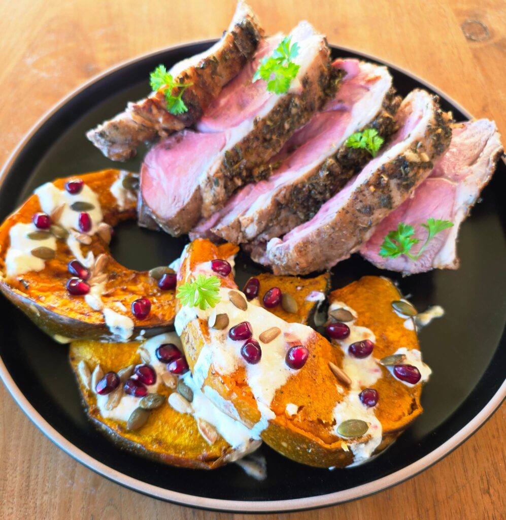 Serving suggestion for lamb roast: slices of lamb roast served alongside pumpkin, pomegranate and lemon garlic yogurt, served on a black plate against a wooden table.