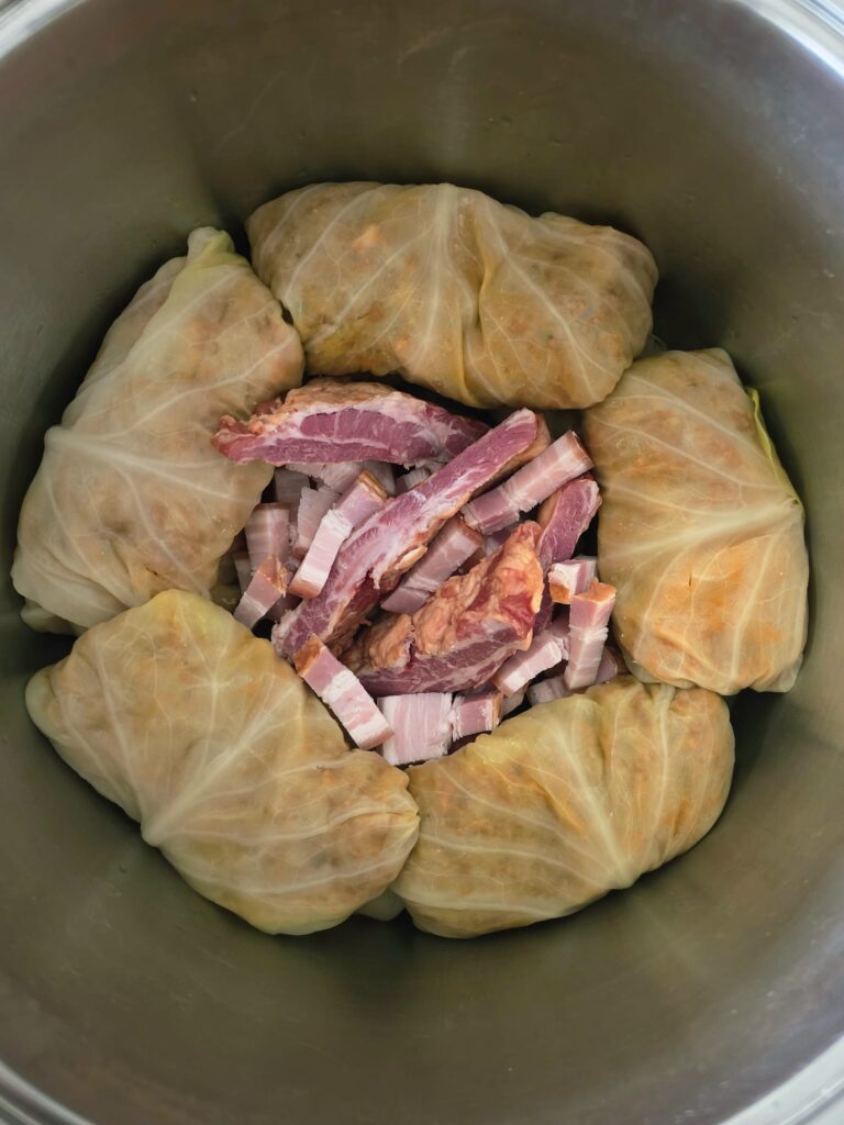 Overhead photo of a large stainless steel pot, showing the Balkan Stuffed Cabbage Leaves (Sarma) arranged on the sides of the pot in a stack, with smoked speck and smoke pork ribs placed in the middle of the stacks of rolls