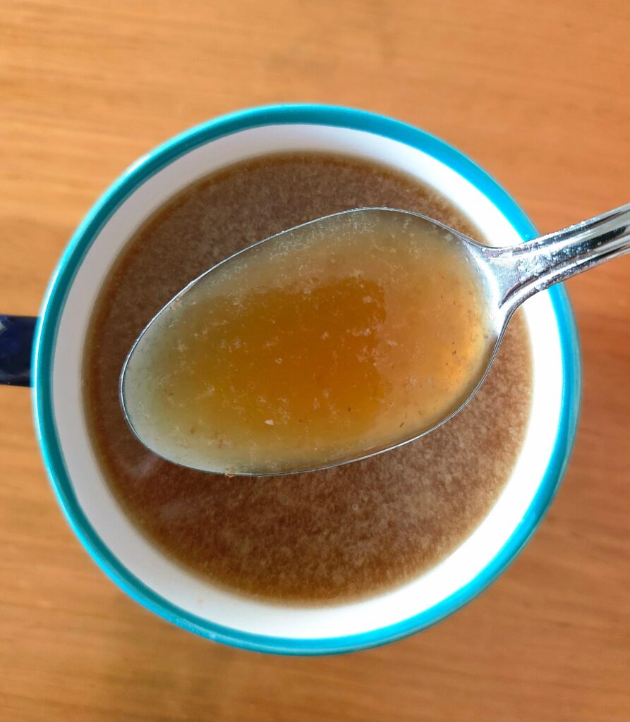 A cup of bone broth set on a wooden table, with a metal spoon lifting some bone broth up to the camera.