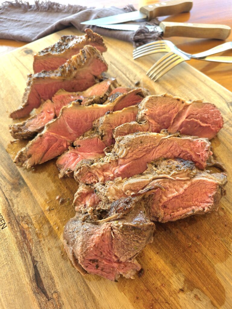 Thick pink slices of this Easy Butterflied Lamb Leg with Sumac, Lemon and Oregano spread out on a wooden board, with knives and forks and a blue tea towel in the background