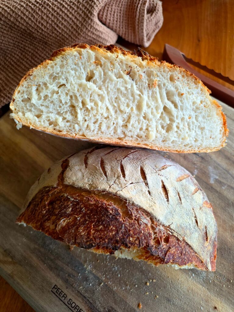 Cross section of a freshly baked loaf of this Easy Beginner No-Knead Sourdough showing it's open, airy crumb, with a bread bow and a brown tea towel in the background