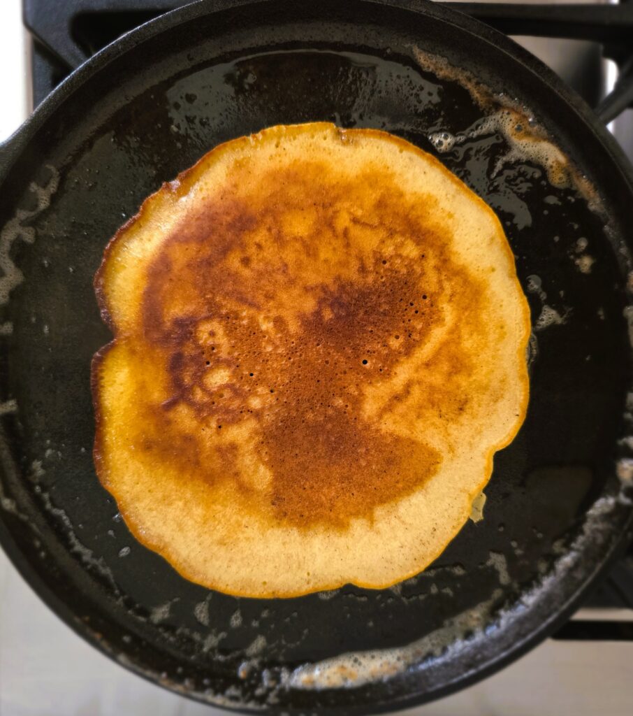 Flipped pancake cooking in cast iron skillet showing the golden brown colour and crispy edges from being cooked in butter